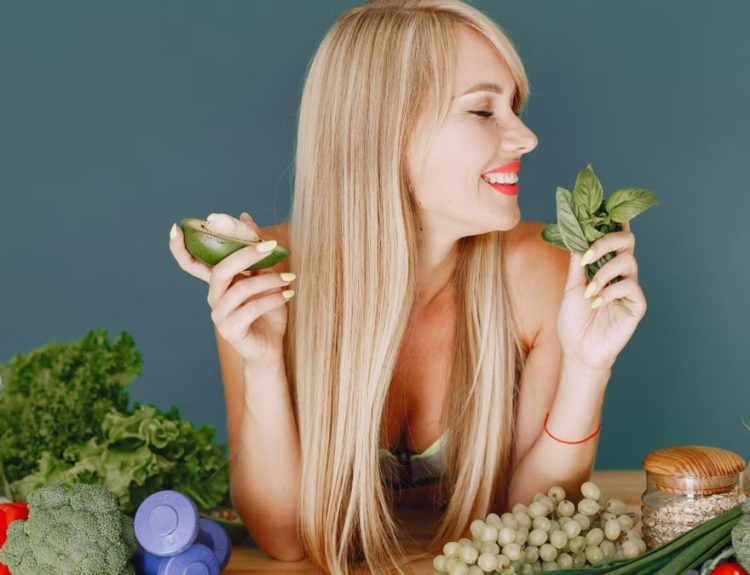 A smiling woman holding avocado and basil, surrounded by fresh vegetables and fruits for healthy eating.